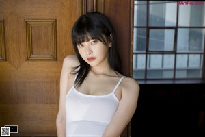 A woman in a white tank top leaning against a curtain.