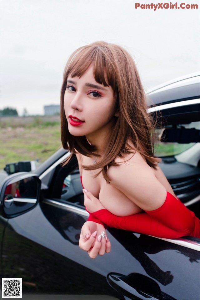 A woman in a red dress leaning on a black car.