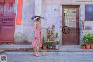 A woman in a pink dress and a straw hat is standing on a sidewalk.