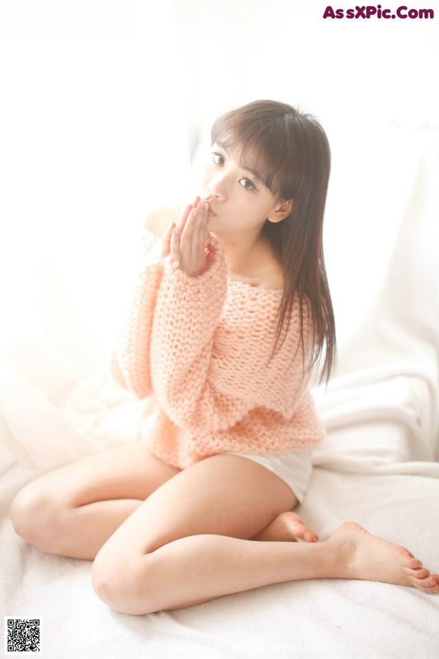 A woman sitting on a bed in a pink sweater.