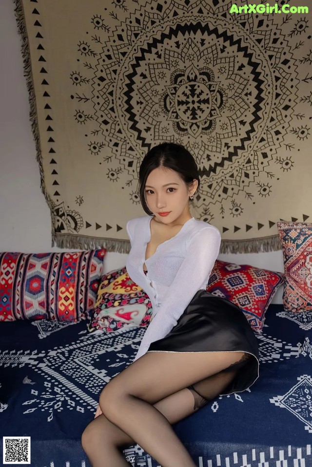A woman sitting on top of a bed in a white shirt and black skirt.